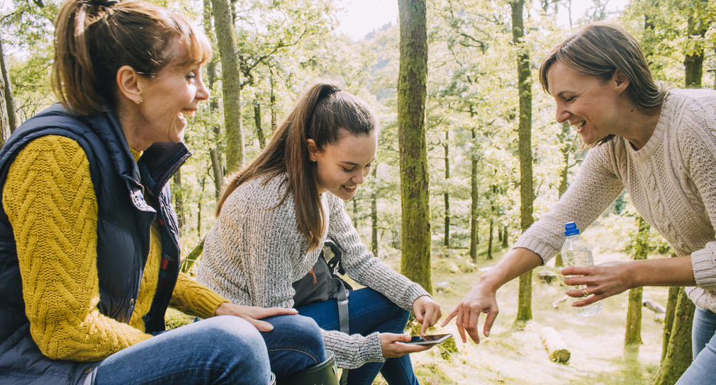 Drei Frauen im Wald. Sie schauen auf ein Smartphone, das die jüngste in der Hand hält.