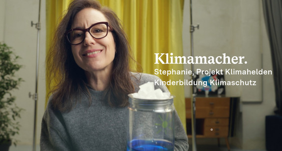 Stephanie Esch schaut in die Kamera, im Hintergrund ist ein Vorhang, im Vordergrund ein Glas mit blauer Flüssigkeit zu sehen.