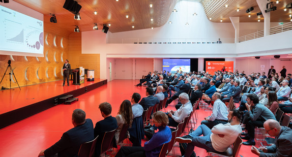 Ein Bild aus einer Veranstaltungshalle, in der viele Personen sitzen und auf eine Bühne schauen. Auf der Bühne steht ein Vortragender, welcher Präsentationsfolien zeigt. Die Zuschauerinnen und Zuschauer sind alle aufmerksam nach vorne gerichtet, um den Vortrag zu verfolgen.