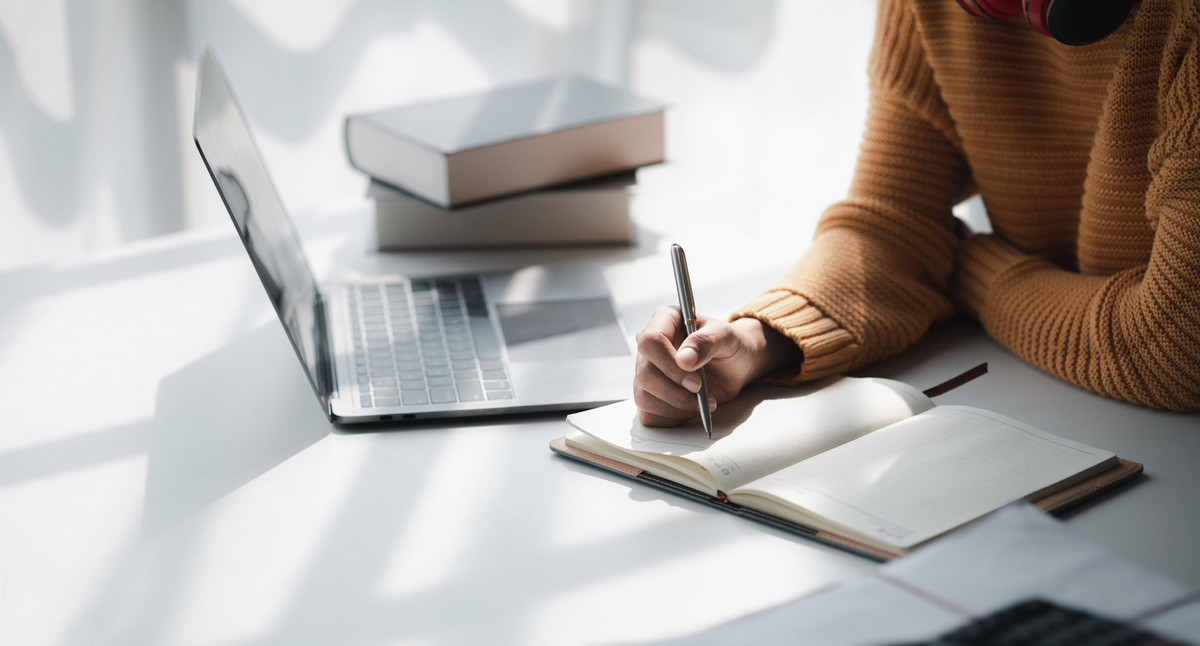 Person schreibt etwas in ein Notizbuch, neben ihr steht ein Notebook und liegen Bücher auf dem Tisch